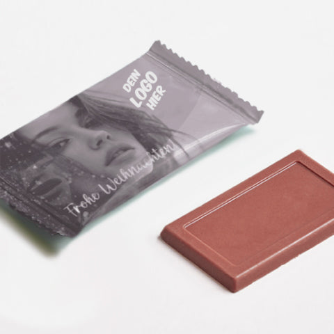 Eine bedruckte Verpackung eines Schokoladentäfelchens, daneben eine kleine Mini-Schokoladentafel. Der Druck der Verpackung zeigt ein Model mit Schneeflocken im leicht wehenden Haar, unten auf der Verpackung steht "Frohe Weihnachten", Platz für ein individuelles Salonlogo in der rechten, oberen Ecke.
